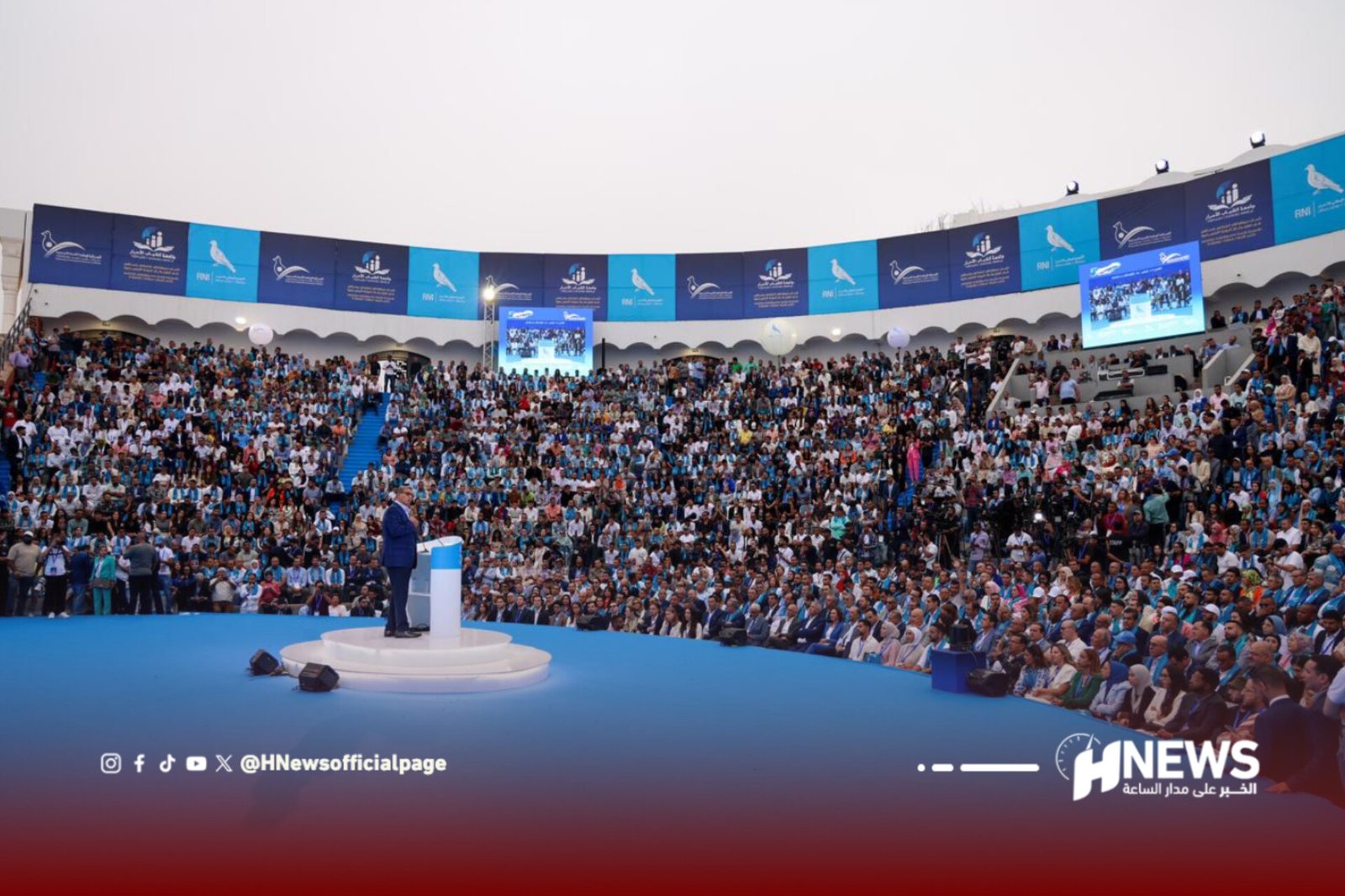 Speaker at a podium addressing a massive stadium crowd under blue banners and big screens on the sides.