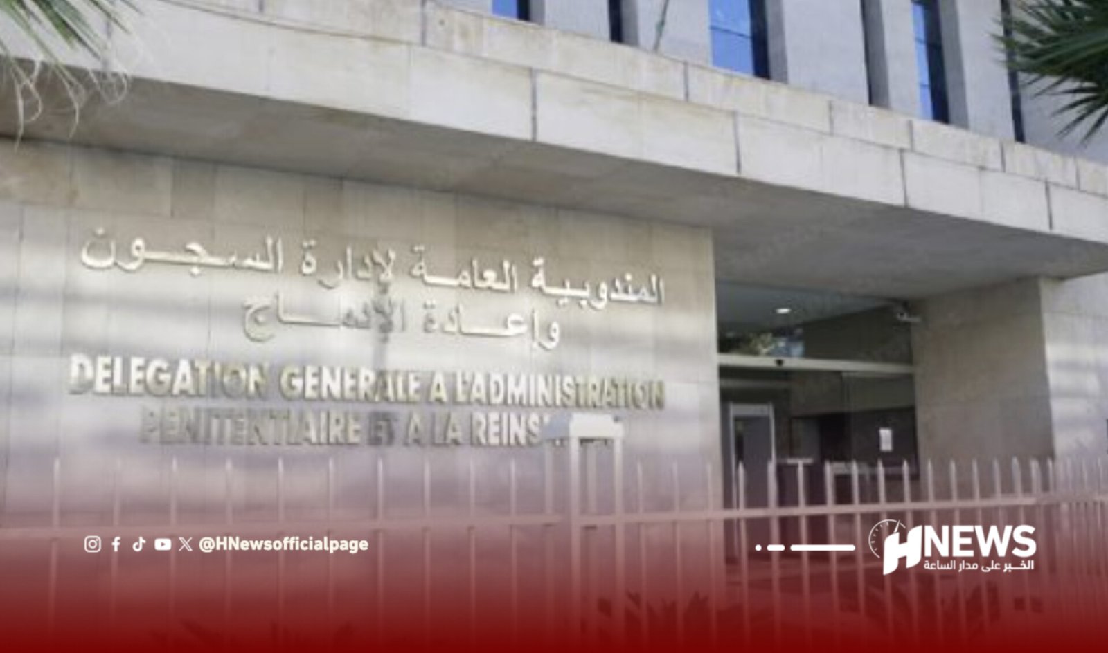 Front entrance of a government administration building with Arabic signage on a stone wall and a metal fence in the foreground.