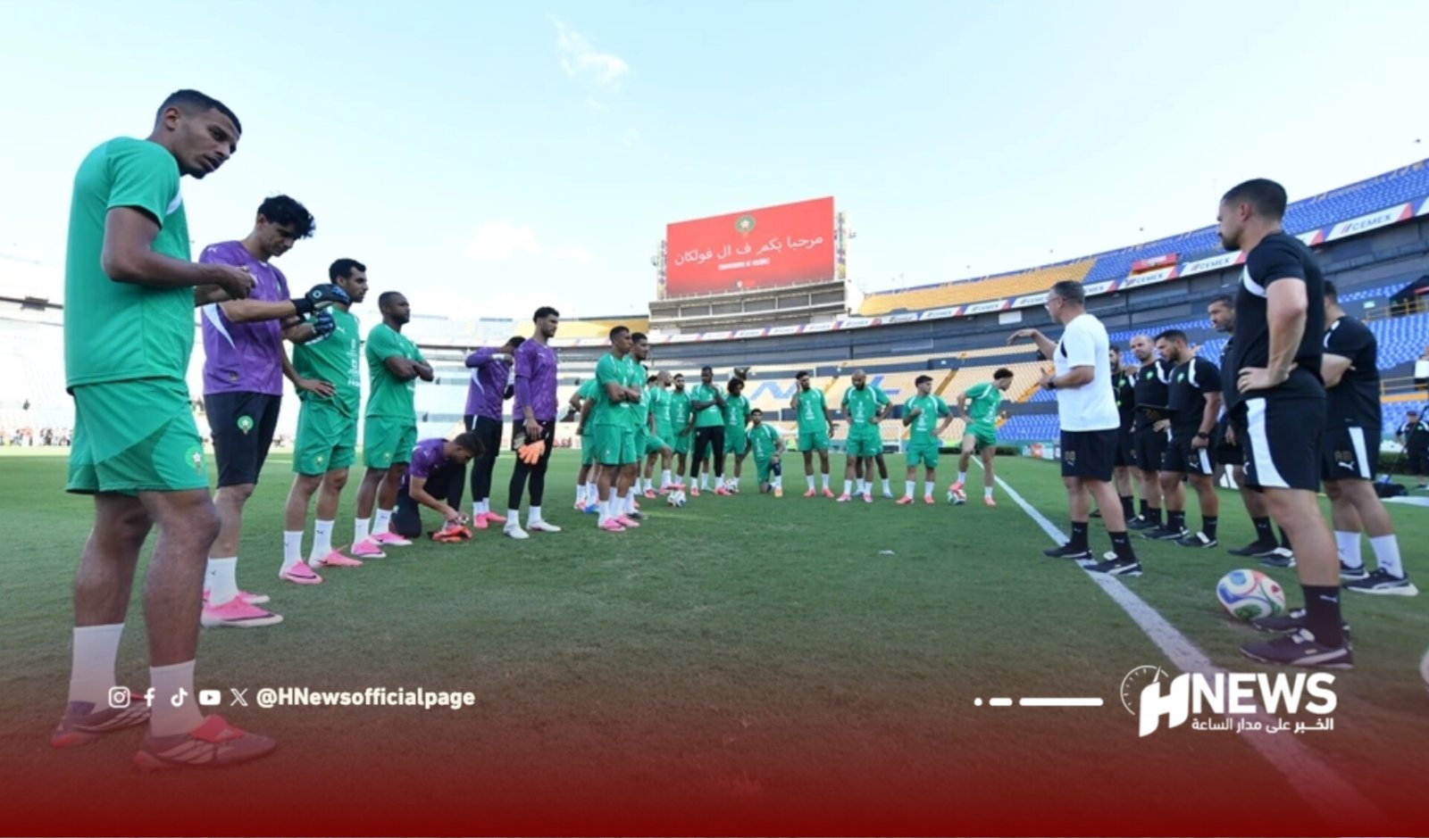 Soccer players in green training kits stand in two lines on a stadium field while a coach talks to them.