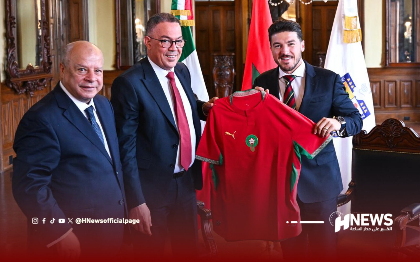 Three men in suits hold a red Moroccan national team jersey during a formal ceremony, with flags in the background behind them.