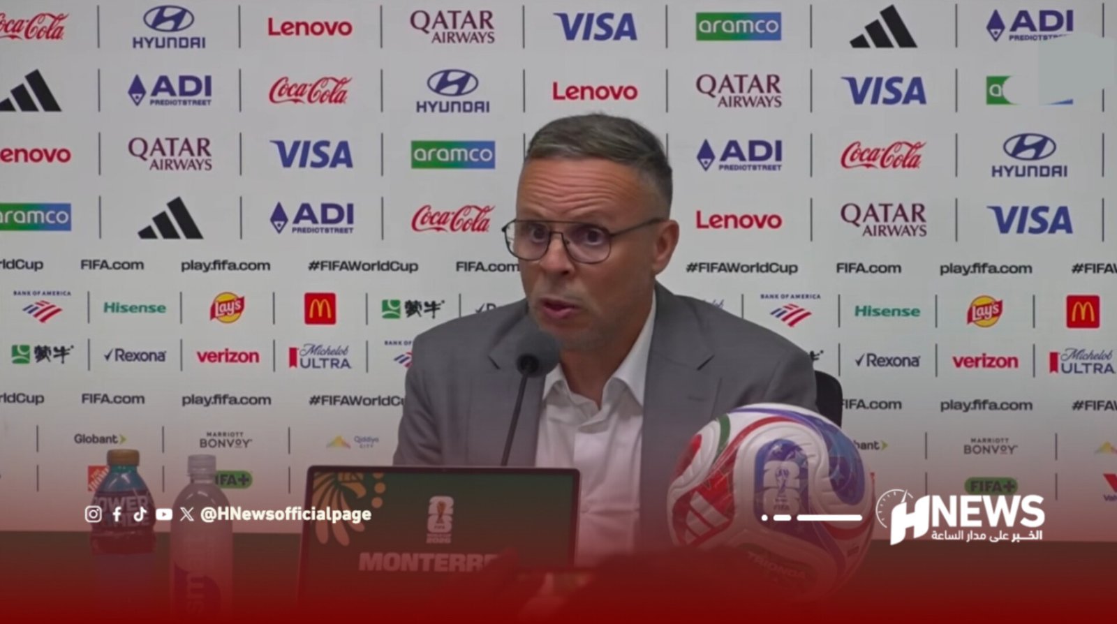 Man in a suit at a press conference table, speaking into microphones with a FIFA World Cup sponsor backdrop and a soccer ball on the table.