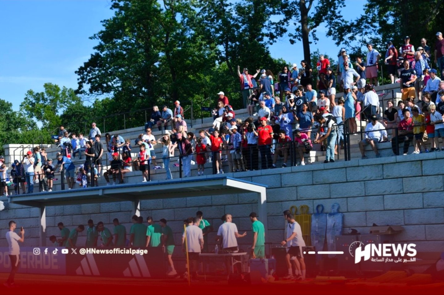 Crowd of spectators on concrete bleachers watching a sports event on a sunny day, with players near a sheltered dugout below the stands.