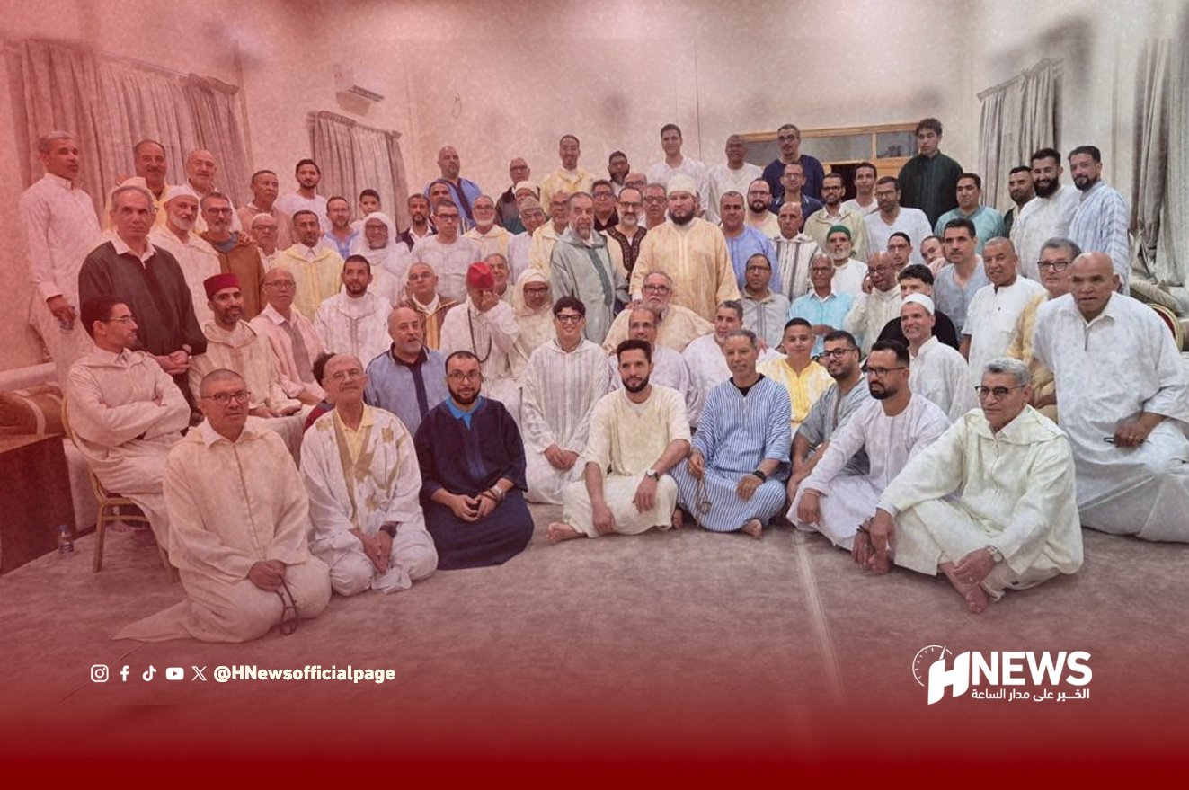 Group of dozens of men in traditional attire posing together in a large room, with a red overlay and HNews watermark at the bottom right.