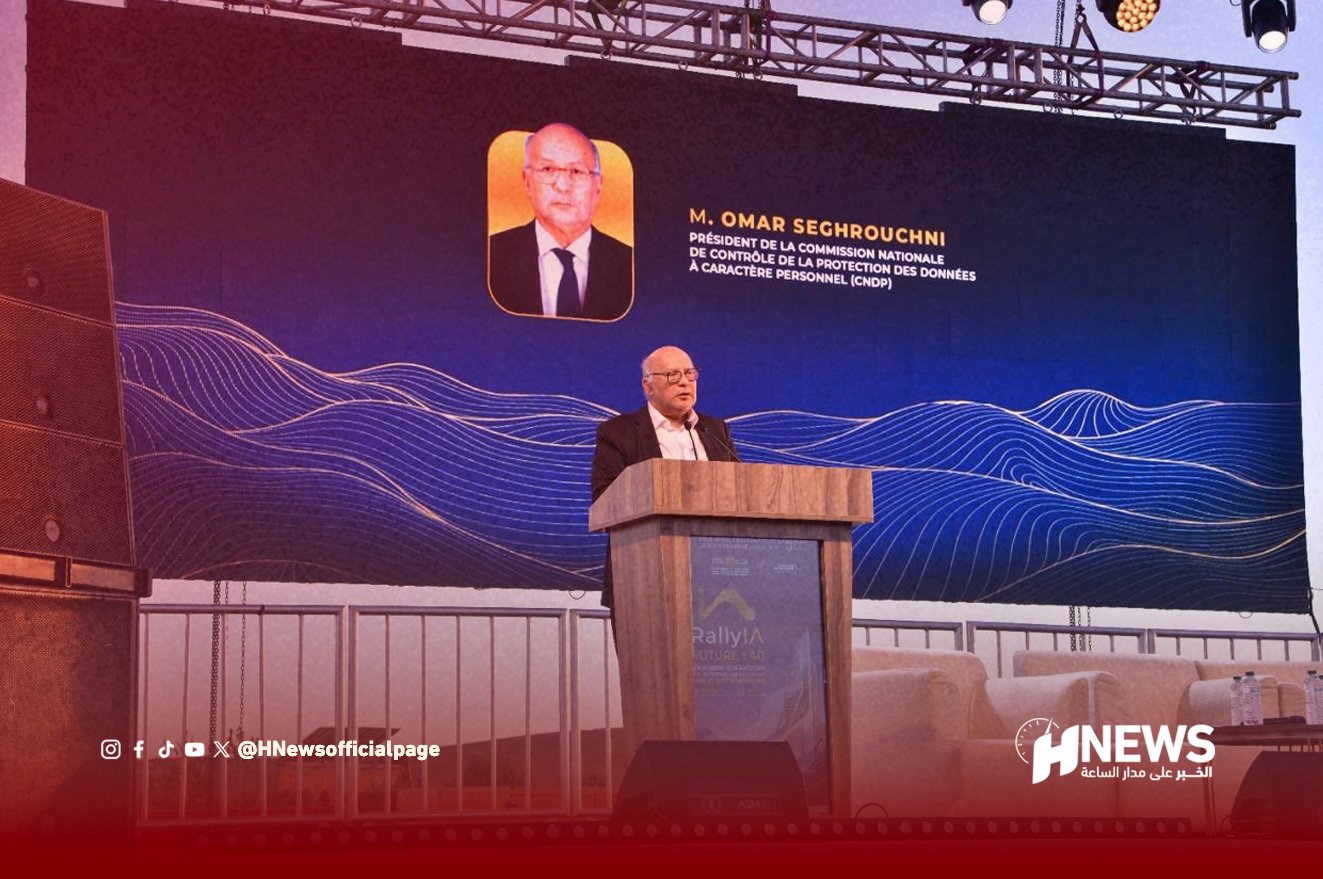 A man in a suit speaks at a wooden podium on a stage, with a large dark blue screen behind him showing a portrait and text.
