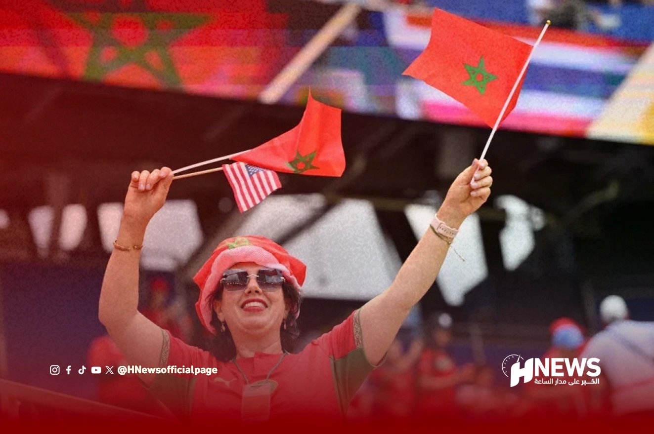Cheerful woman in red waving Moroccan and American flags at a stadium, wearing sunglasses and a red headscarf.