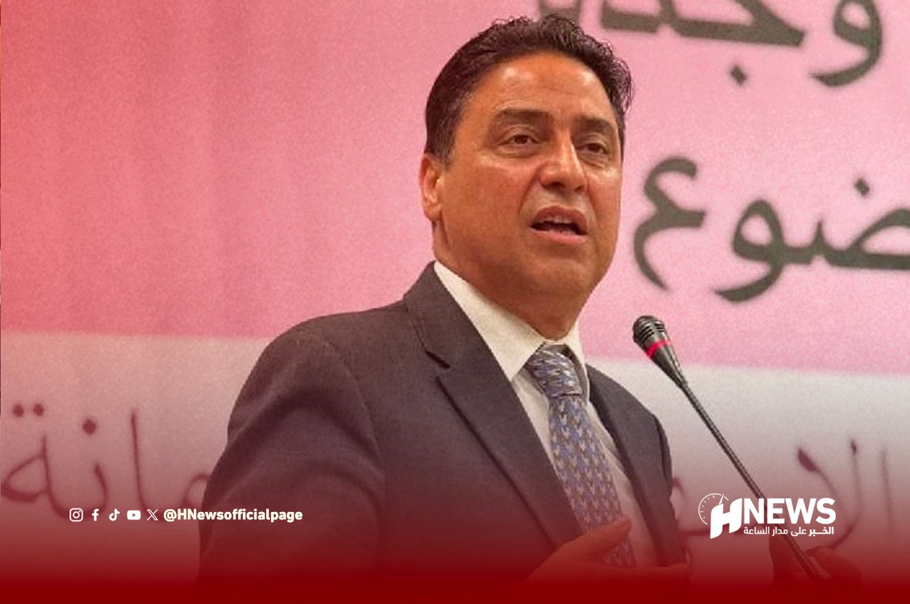 Man in a dark suit and patterned tie speaks at a microphone on a stage with a pink backdrop and a news outlet watermark in the corner.