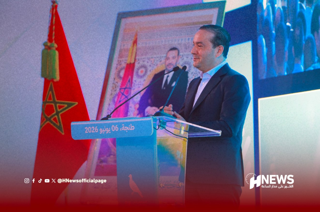 Man in a suit speaks at a clear podium during a formal event; Moroccan flag and a framed portrait are in the background (Tangier, 06 June 2026).