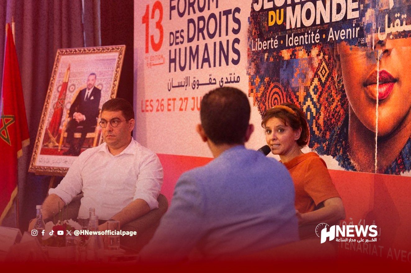 Panel discussion at Forum des Droits Humains with a woman speaking into a microphone; two men sit at a table in front of a large colorful banner.