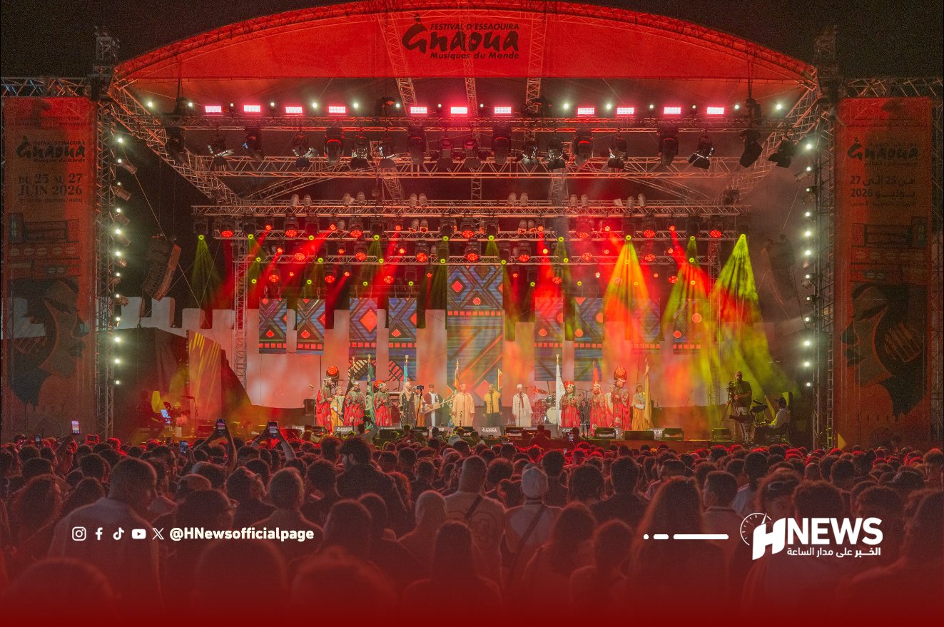 Outdoor night concert on a large stage with colorful red and yellow lights and a dense crowd watching performers in traditional outfits at the Gnaoua festival surface.