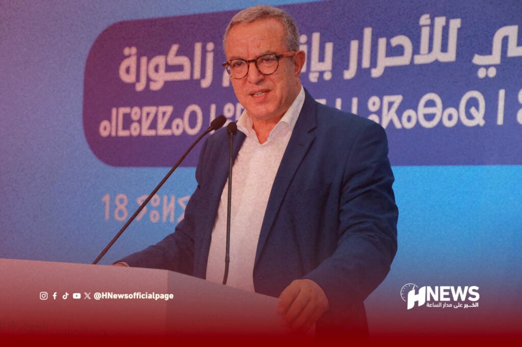 الأحرار أوجار | H-NEWS آش نيوز Male presenter in a blue suit and glasses speaks at a podium with two microphones, against a blue/purple backdrop and HNews logo in the corner.
