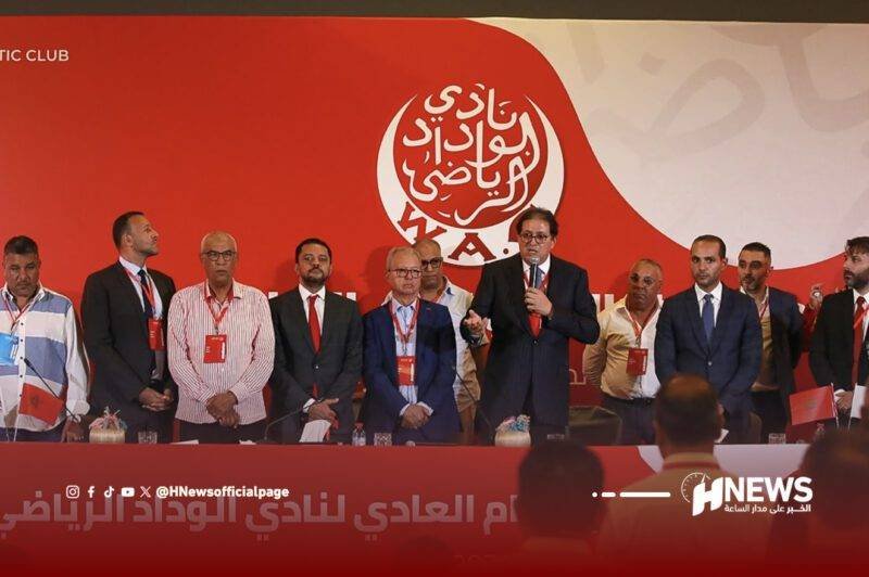 جمع-عام-الوداد-scaled | H-NEWS آش نيوز Group of men in suits standing on a red stage behind a long table, with one man speaking into a microphone in the center and a large circular Arabic logo on the red backdrop behind them.