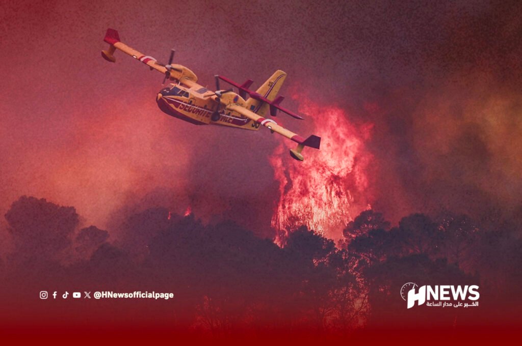 حرائق فرنسا | H-NEWS آش نيوز Yellow air tanker plane flying above a forest wildfire, spraying fire retardant against orange-red flames and smoke.