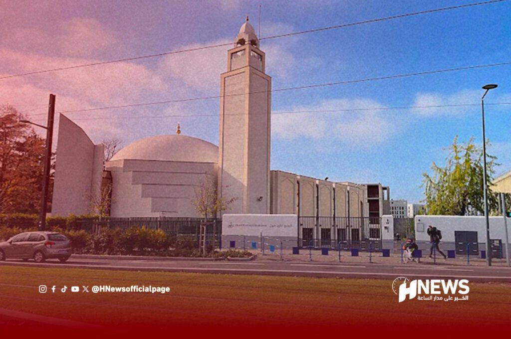 مسجد ليون | H-NEWS آش نيوز Modern mosque with a large dome and a tall minaret along a street, with a car parked and pedestrians nearby; HNews watermark bottom-right.
