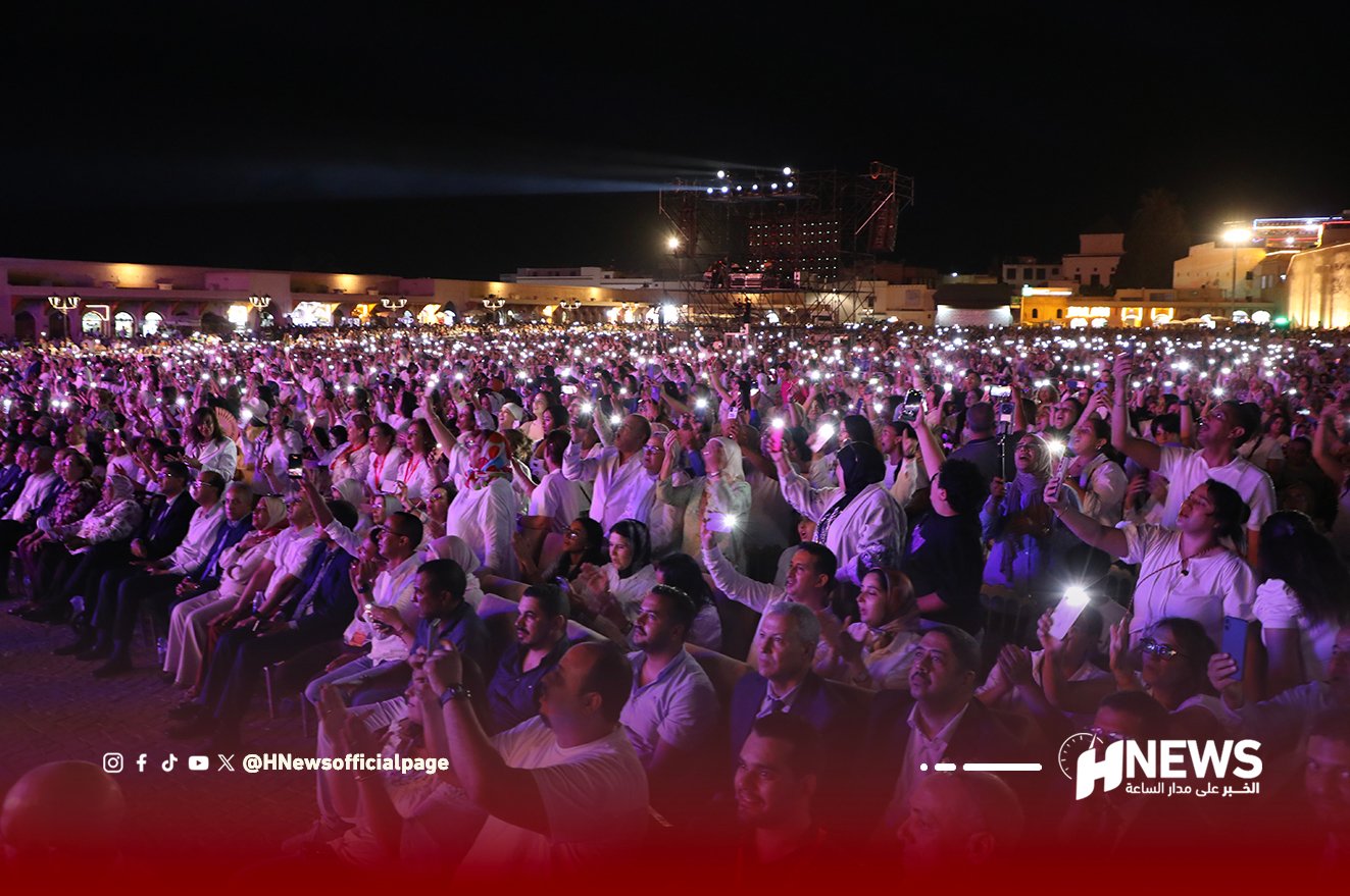 مهرجان عيساوة 3 | H-NEWS آش نيوز Large nighttime crowd at an outdoor event, many people holding up illuminated phone screens toward a distant stage with lights and a news logo in the corner