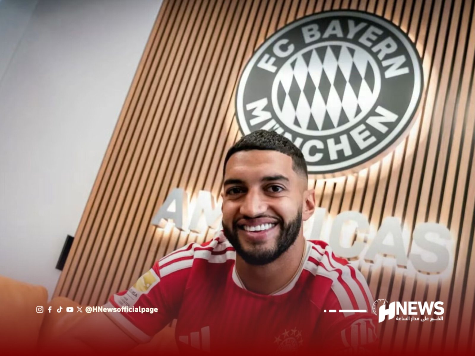 Smiling man in a red Bayern Munich jersey takes a selfie in front of a wall with the FC Bayern logo behind him.