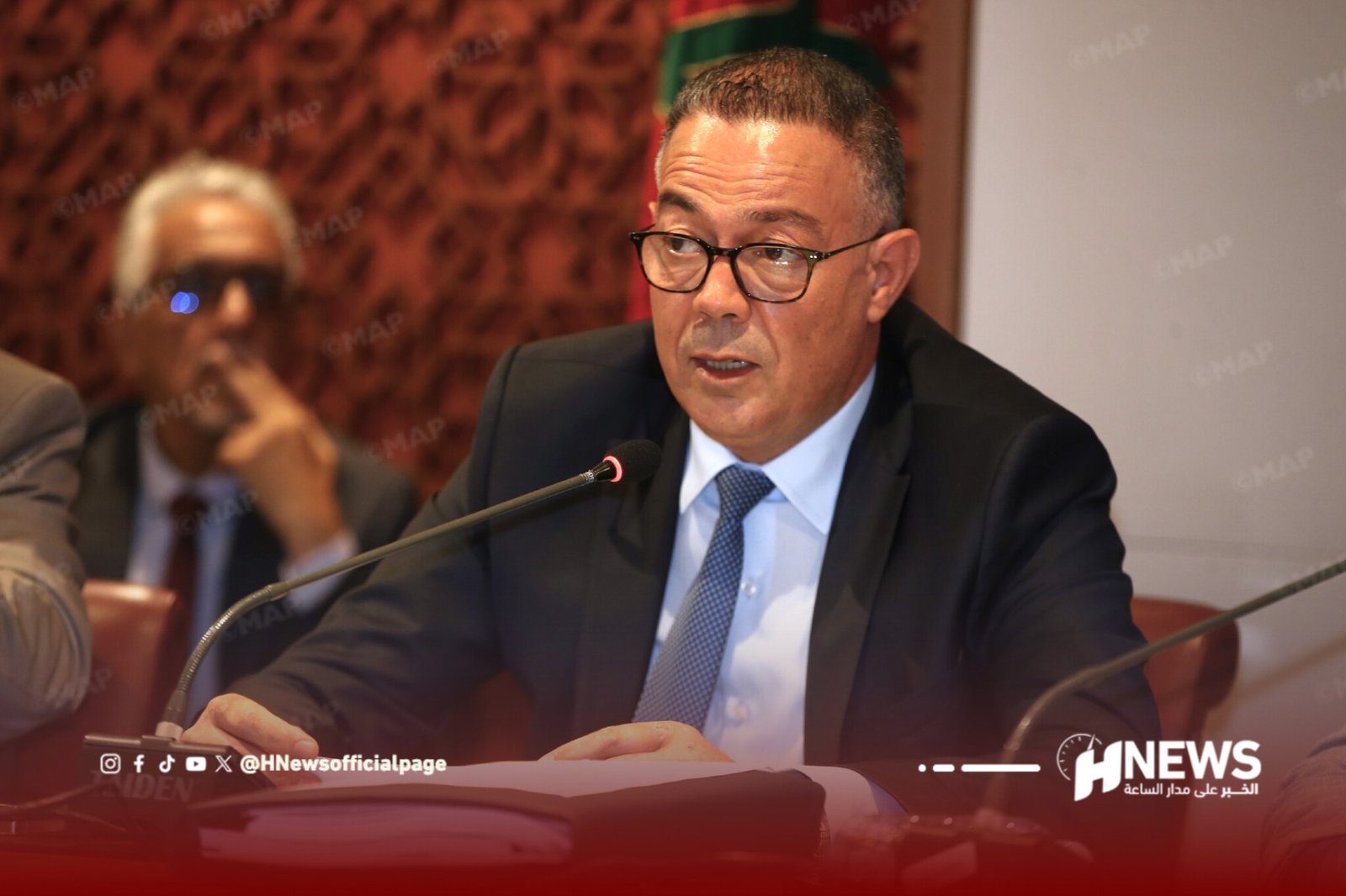 Middle-aged man in a dark suit and glasses speaks at a panel, microphone in front, blurred audience behind him.