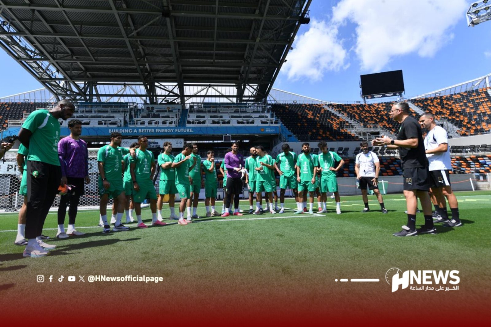 Soccer players in green uniforms stand in a line on the field while a few coaches speak to them in a sunny stadium.