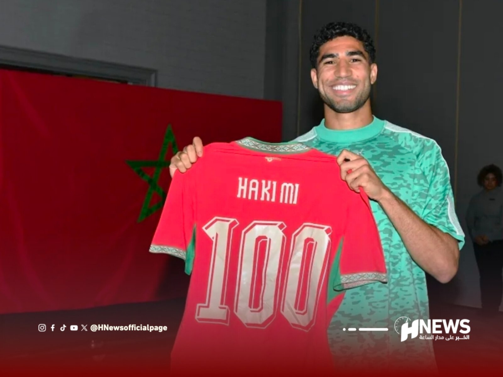 Man smiling while holding a red sports jersey with white outlined number 100; Moroccan flag in background.