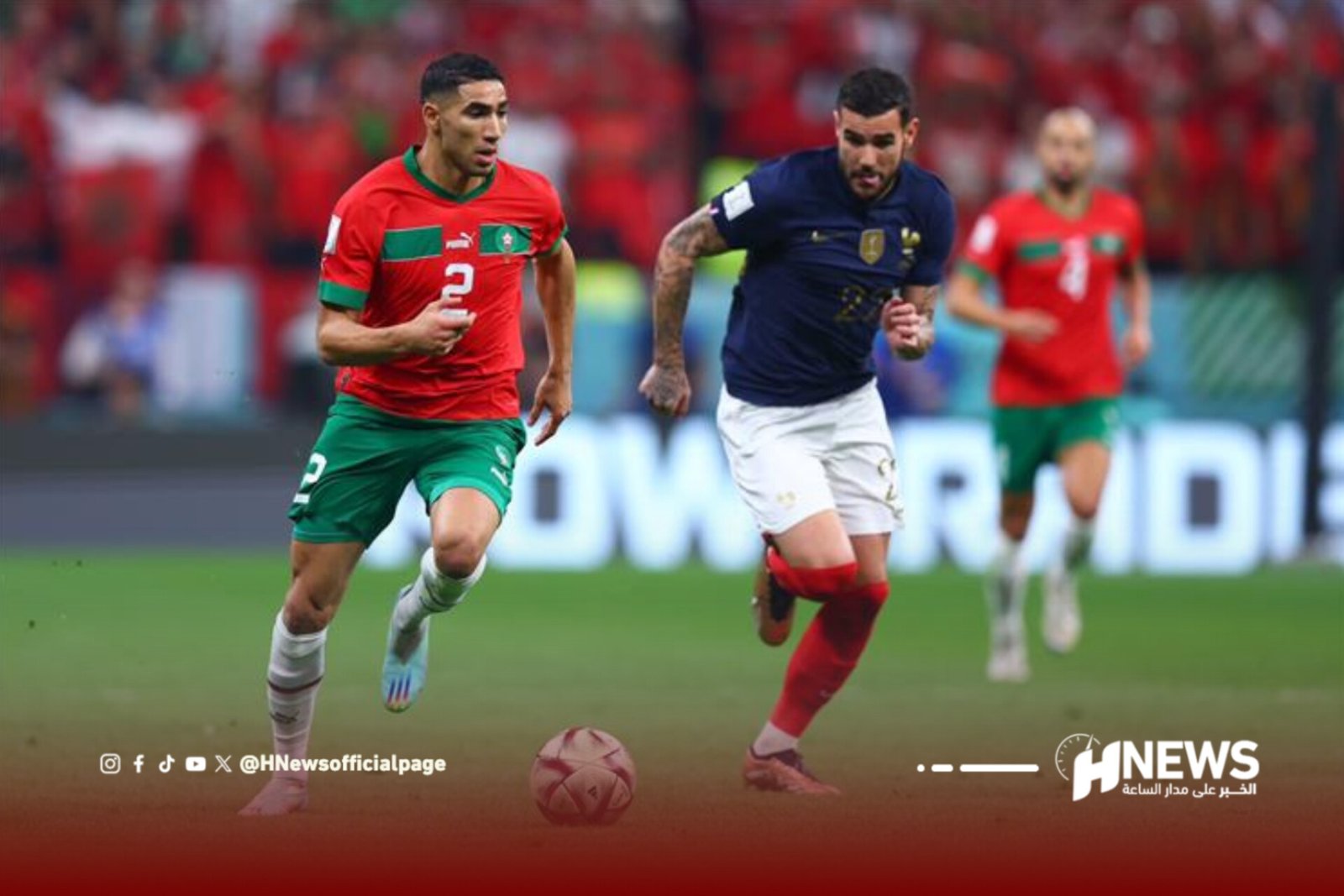 Two soccer players chase the ball on a green field during a match; one wearing red-green kit, the other in navy, with a red-clad crowd behind. Note: HNews watermark visible bottom right.