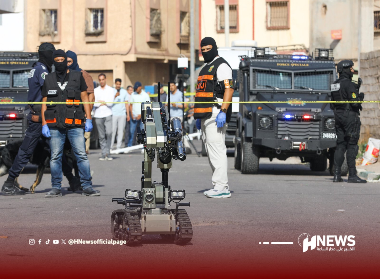 Remote-controlled military robot on a street with masked guards and armored vehicles in the background at a security drill or operation.