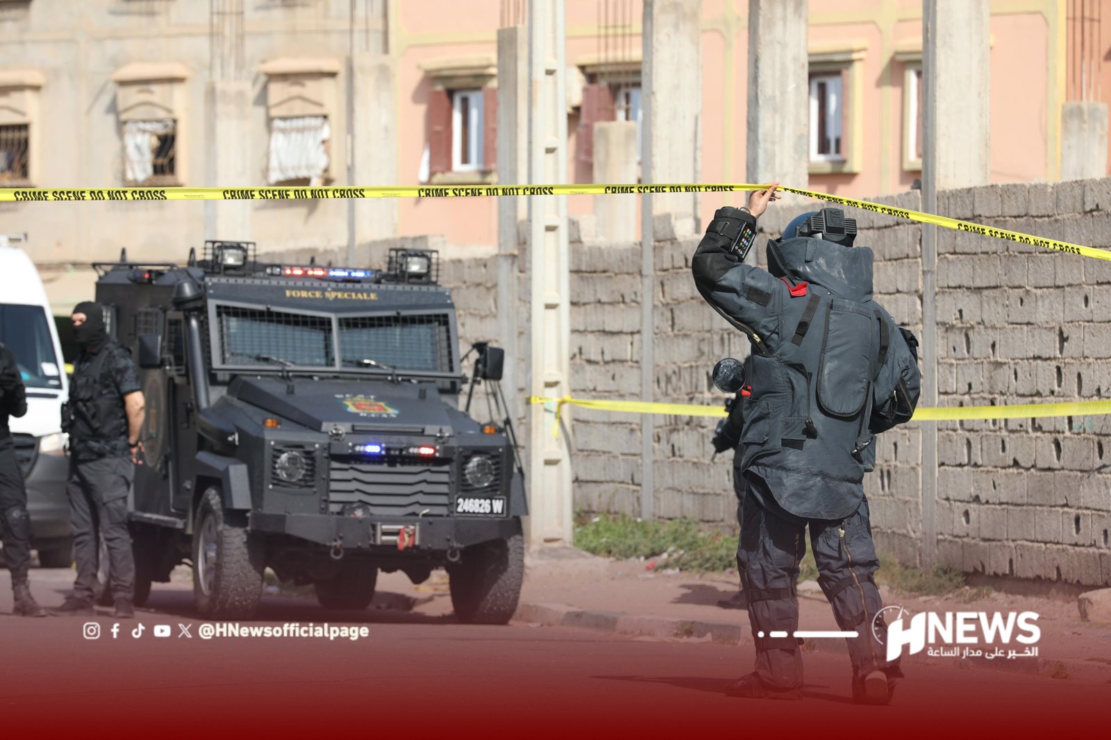 InShot_20260706_131253965 | H-NEWS آش نيوز Armed security officer in body armor places yellow'Crime Scene Do Not Cross' tape across a street, with an armored vehicle in the background.