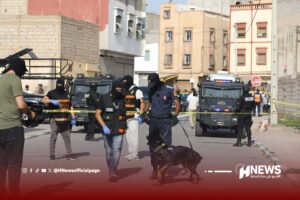 Police and armed officers in tactical gear guard a cordoned urban street with armored vehicles and a police dog nearby, as reporters and bystanders watch behind yellow crime-scene tape.