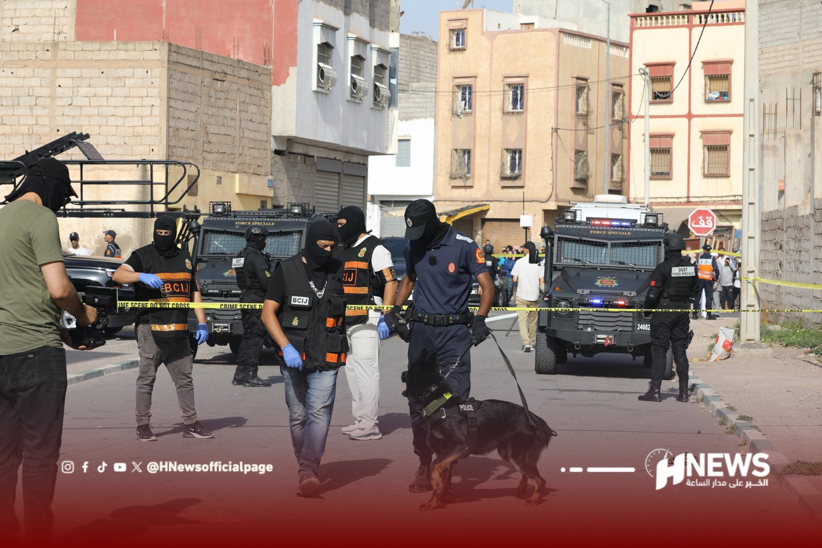 InShot_20260706_131314981 | H-NEWS آش نيوز Police and armed officers in tactical gear guard a cordoned urban street with armored vehicles and a police dog nearby, as reporters and bystanders watch behind yellow crime-scene tape.