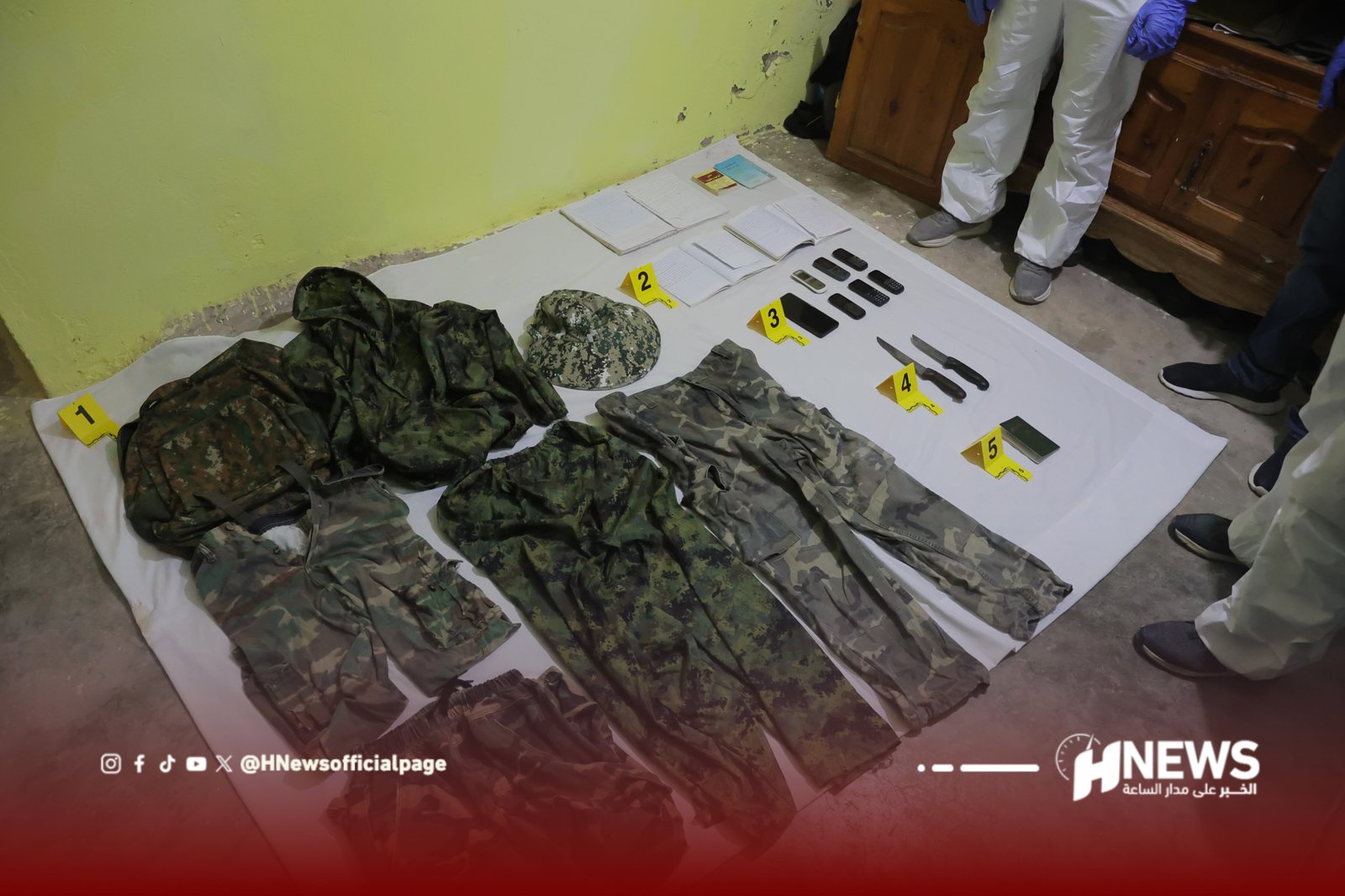 InShot_20260706_131449334 | H-NEWS آش نيوز Military camouflage uniforms and gear laid on a white sheet with five labeled evidence markers (1–5), plus notebooks and multiple phones; people in protective suits stand nearby.