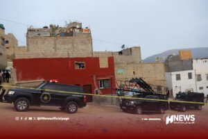 Emergency scene in a dense urban area: a red building with a partially finished upper level, crime-scene tape cordons off the area, and police vehicles labeled 'Force Speciale' are parked; a few people stand to the left, with mountains in the distance.