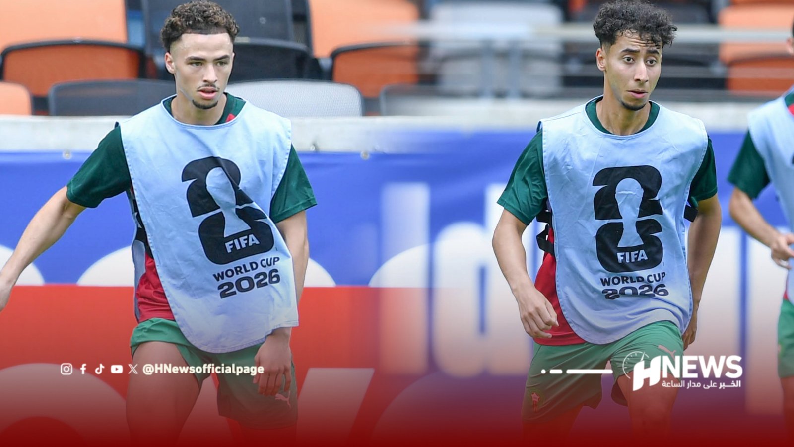 Two male soccer players in green kit wear light-blue bibs with the FIFA World Cup 2026 logo while running on a stadium pitch during practice.