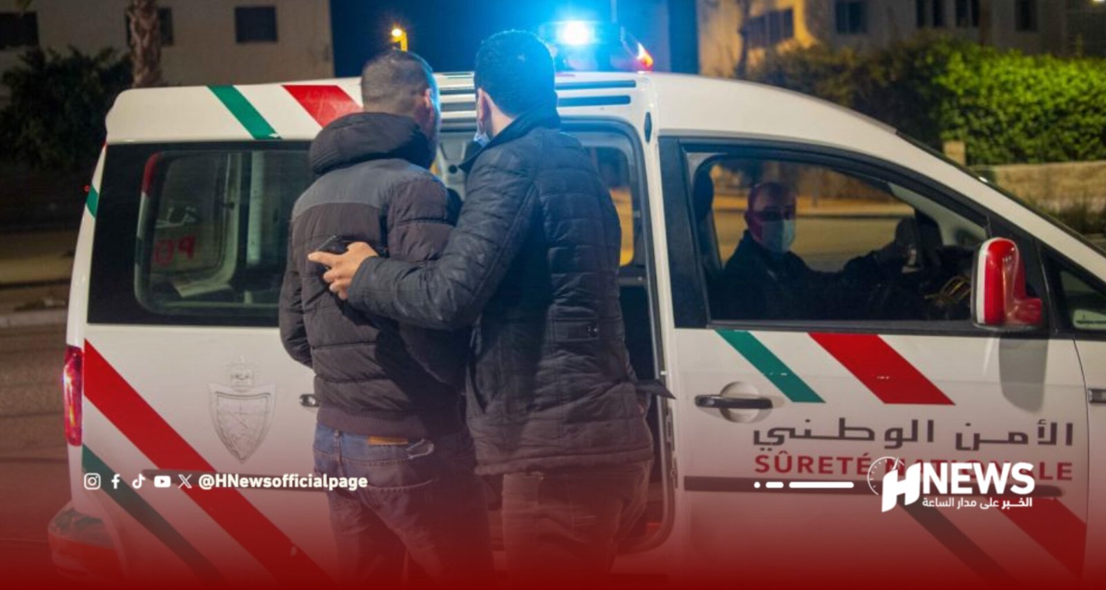 Two men in jackets stand beside a white emergency van with blue lights at night.