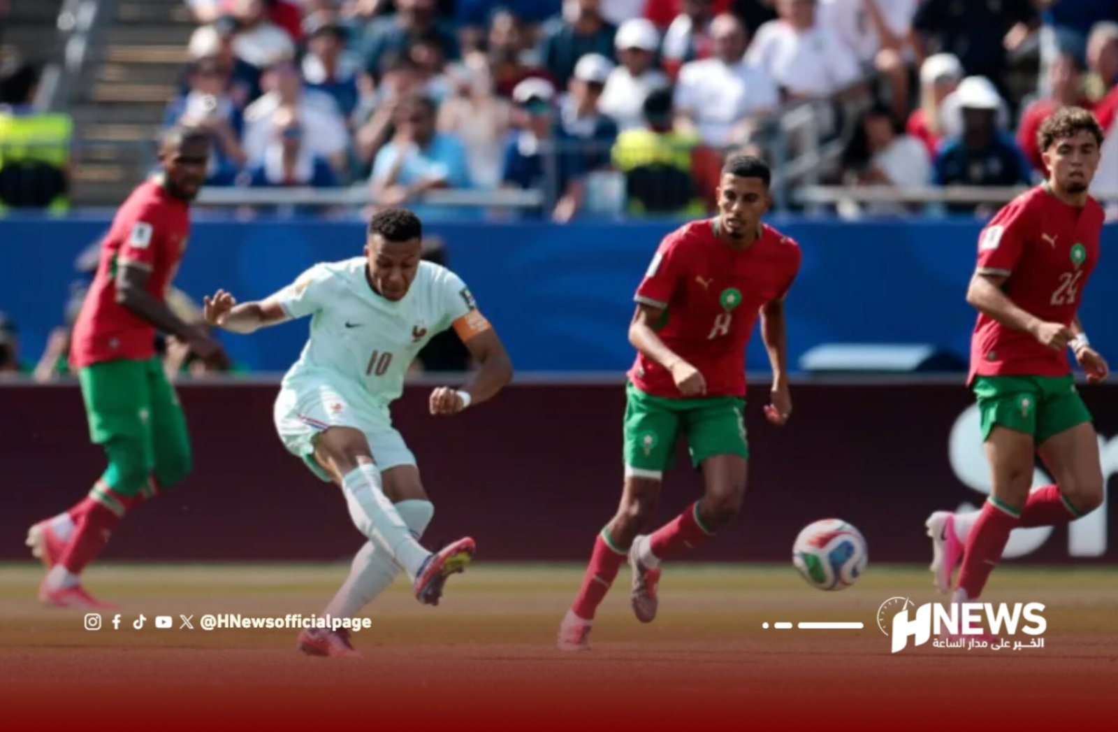 Soccer match with players in red and white uniforms chasing the ball on a sunny field; crowd in stands in the background.