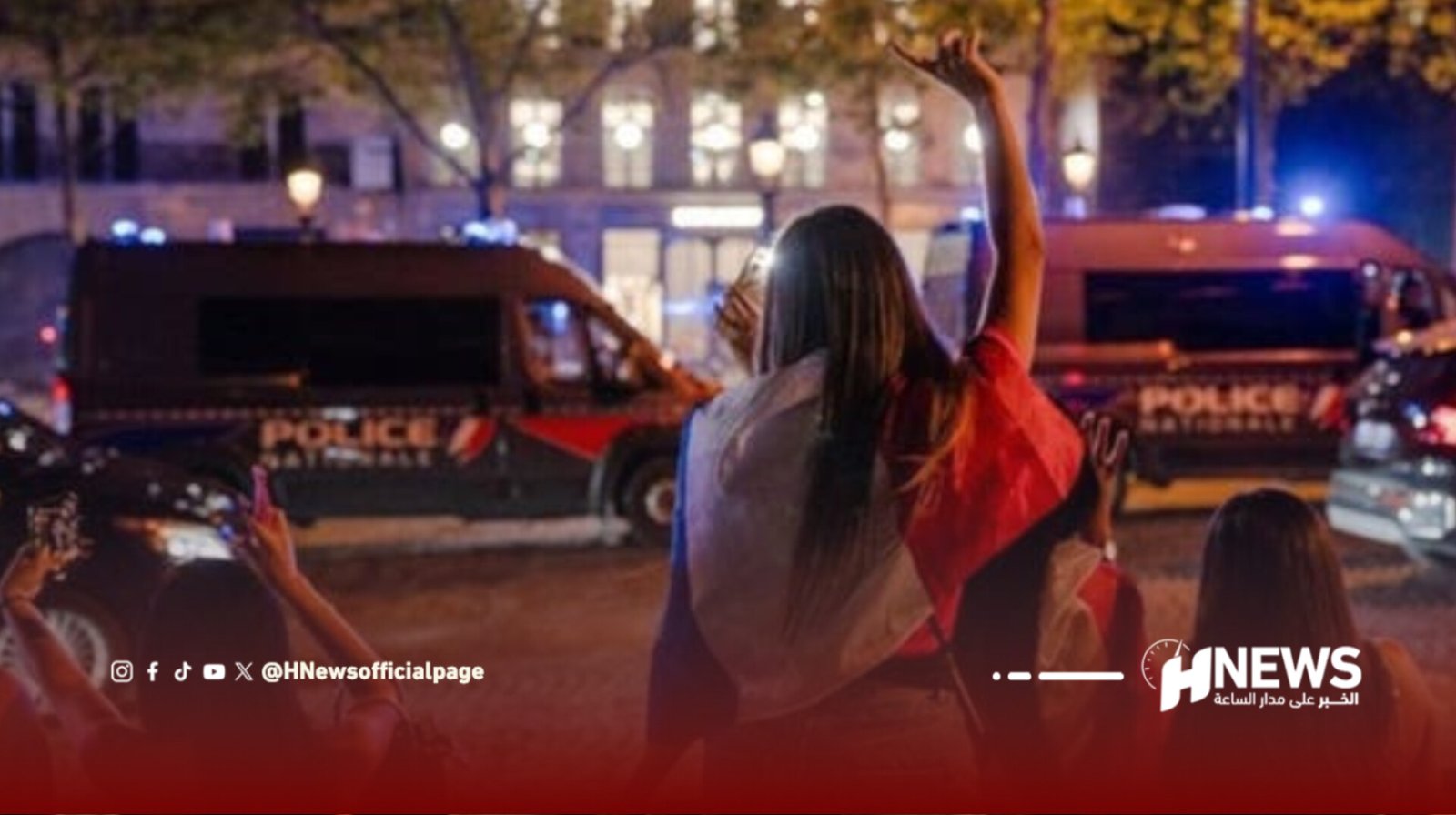 Crowd of protesters raising arms in front of police vans with flashing lights at night in an urban square?scene shows nighttime demonstration with police presence and on-screen news branding.
