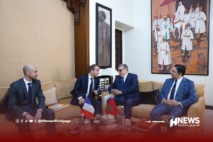 Group of four men in dark suits seated on a beige sofa in a formal meeting room, small Moroccan and French flags on the table in front of them.