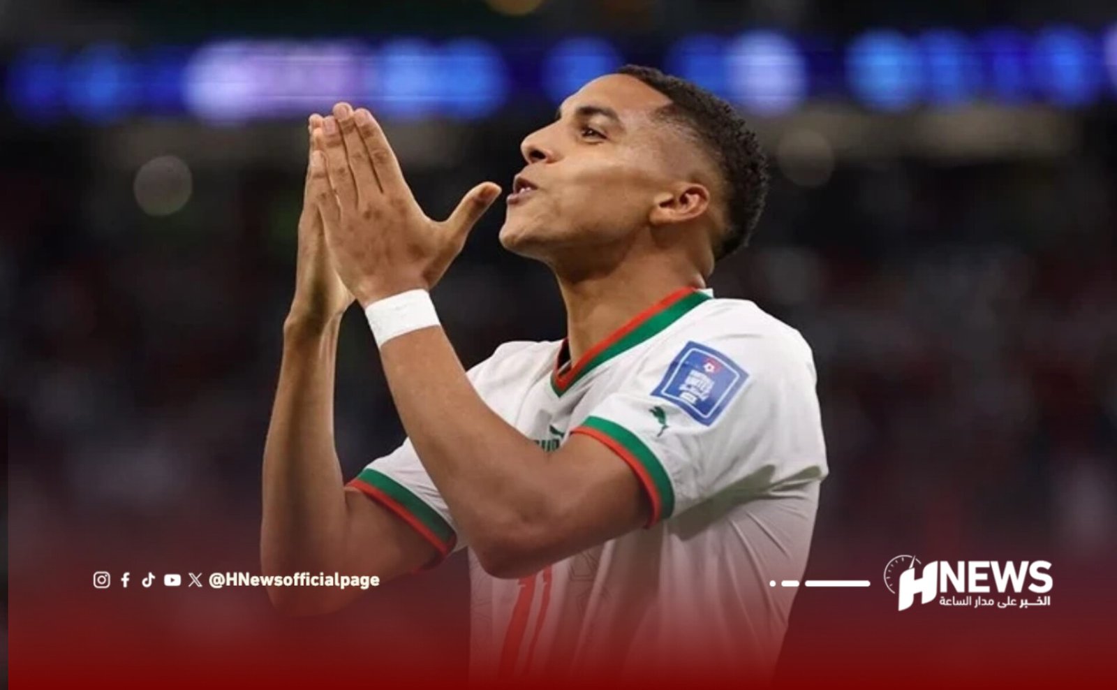 Soccer player in a white jersey with green and red trim prays with hands folded near face on a blurred stadium backdrop.