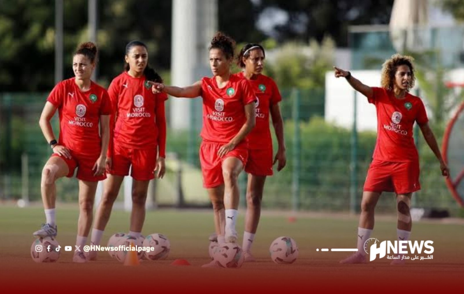 Women’s soccer team in red uniforms standing with soccer balls on a practice field, one player points ahead as others watch.
