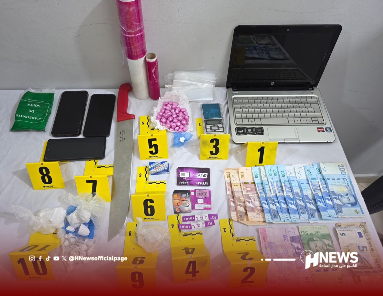 Law-enforcement evidence display on a table: yellow numbered markers surround cash, pills, knives, bags, phones, and a laptop.