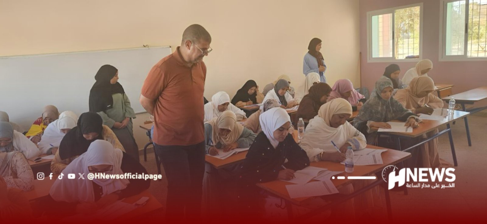 Teacher supervising a classroom as students in hijabs write at their desks during a test or activity, bright windowed room on the right side.