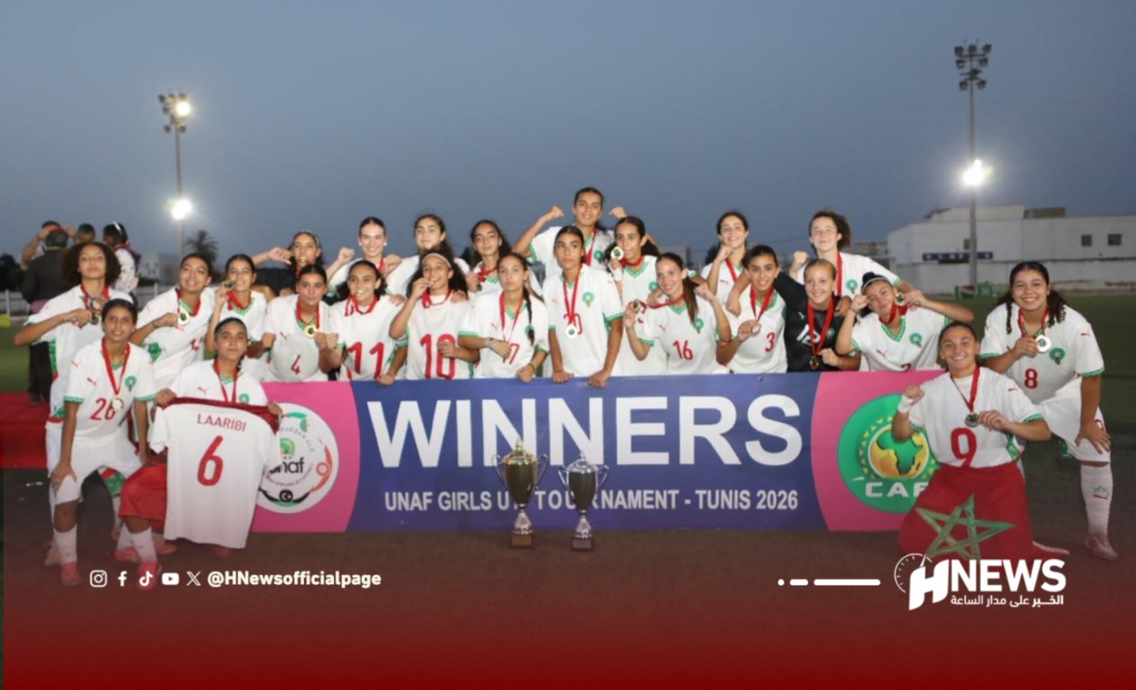 Group of smiling young women soccer players posing with medals in front of a 'WINNERS' banner and trophies on a field.