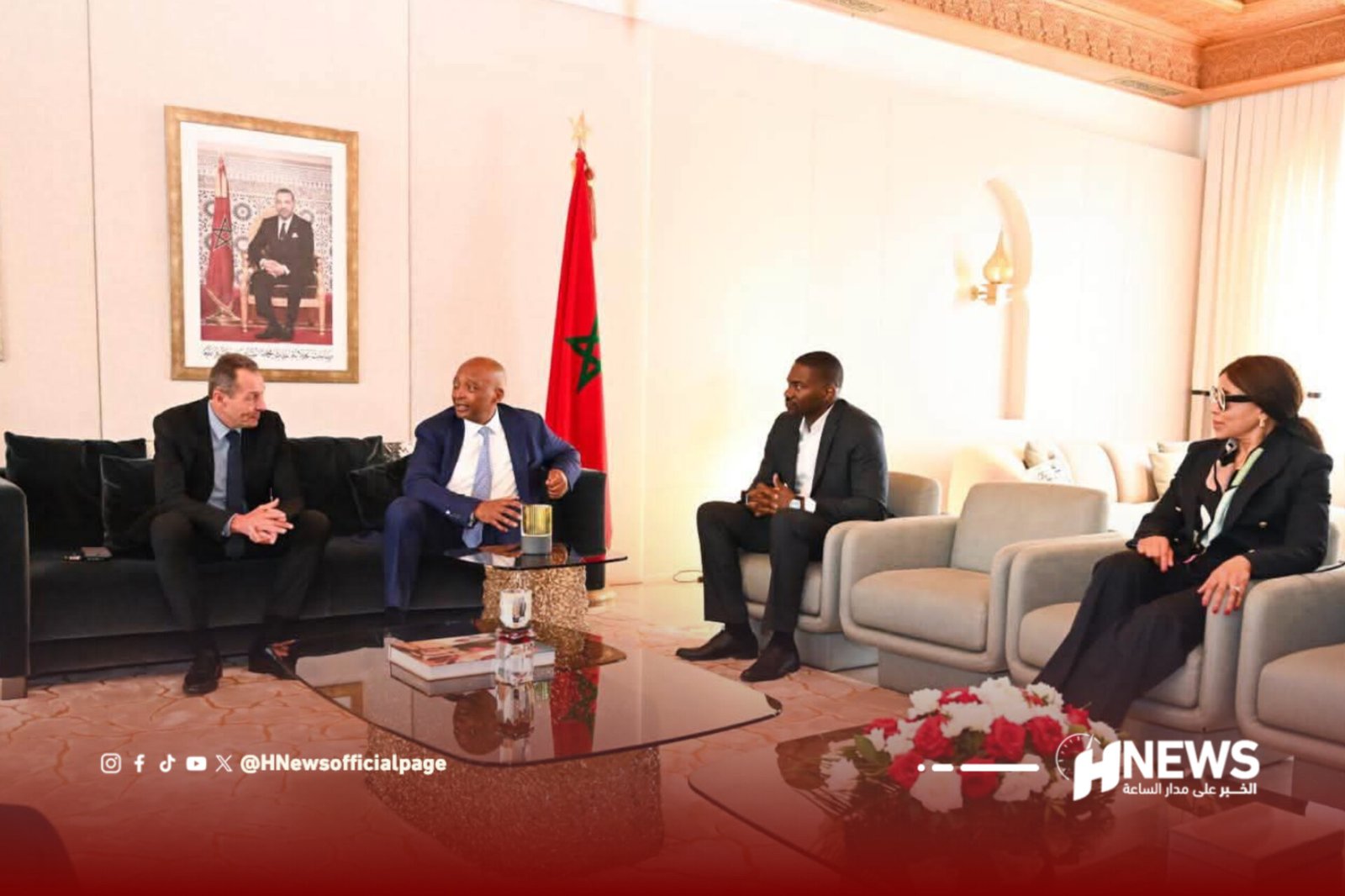 Four adults in a formal meeting in a bright lounge, Moroccan flag and portrait in the background.