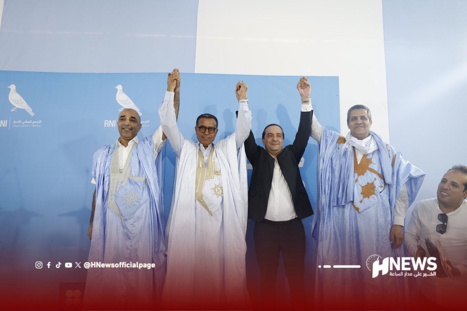 Group of men in blue traditional robes lift their clasped hands in victory on a stage with a blue backdrop and logos behind them.