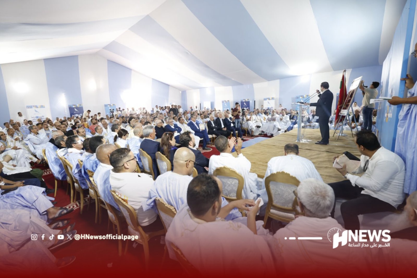 InShot_20260726_154844070 | H-NEWS آش نيوز Large audience seated in a blue-and-white tent watches a man speaking at a podium on stage. (Informative event photo)