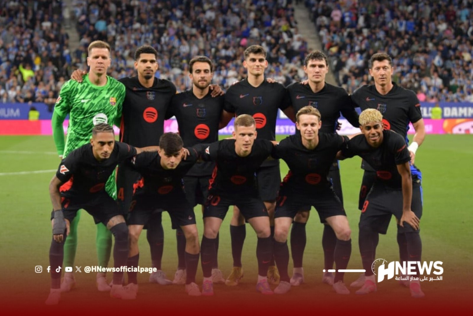 Soccer team posing for a group photo on the field, goalkeeper in green, with fans in the stands behind them.