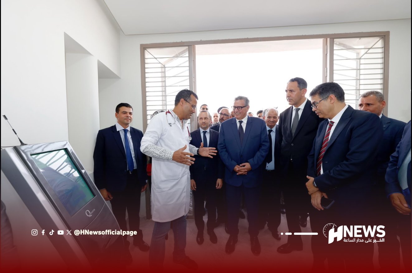 أخنوش مستشفى 4 | H-NEWS آش نيوز Officials in suits listen to a doctor in a white coat explaining a medical kiosk in a bright room with a windowed door visible behind them.