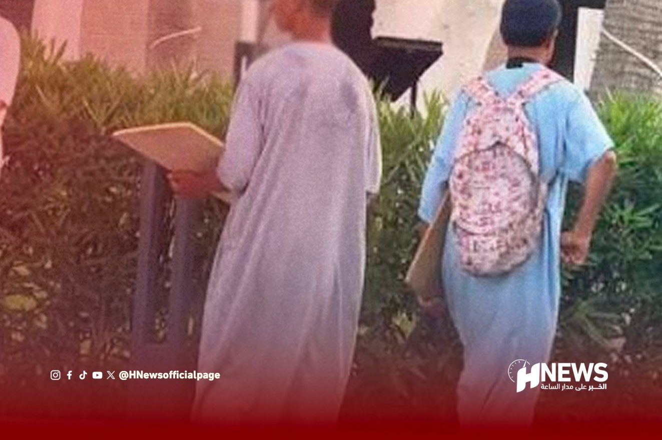 Two people walk away along a hedge; one in a gray robe, the other in blue with a pink floral backpack; HNews watermark.