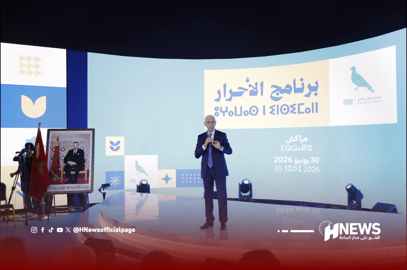 Male speaker in a blue suit delivers a talk on a curved stage with a large digital screen behind him at a conference, flags and a framed portrait to the left.