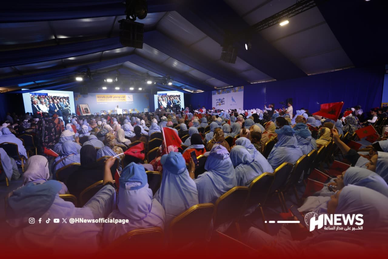 تجمع الأحرار بالداخلة | H-NEWS آش نيوز Large conference hall filled with people seated facing a stage, many wearing blue headscarves, some waving red flags; banners and screens on stage.