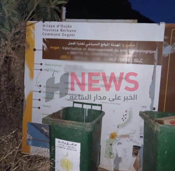 مغارة زكزل | H-NEWS آش نيوز Outdoor project signboard with Arabic and French text about the development of a speleological site; two green trash bins sit in front.