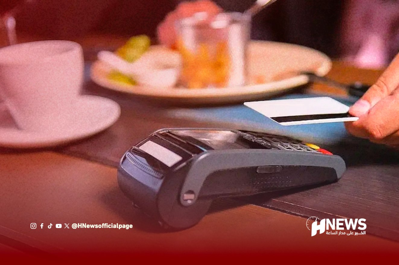 Card payment terminal on a wooden table with a hand holding a card ready to swipe; plate and cup in the background (credit card reader scene)