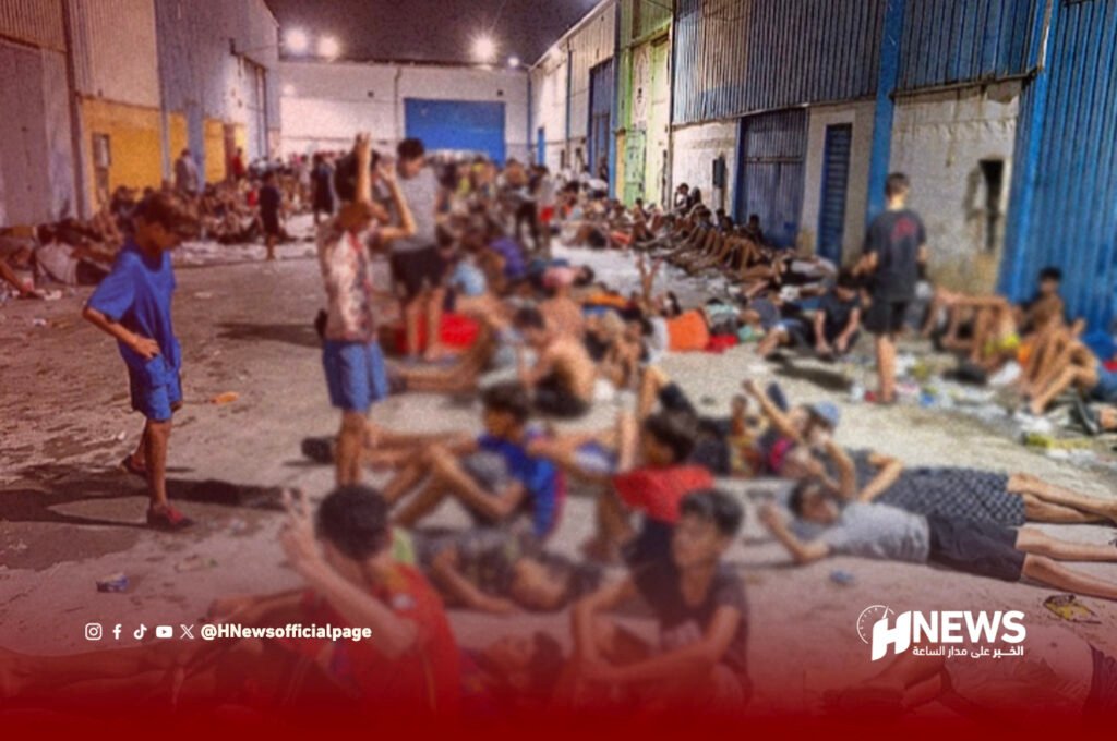 Crowded indoor shelter with people sitting and lying on the floor along a long hallway, appearing tired in a humanitarian setting.